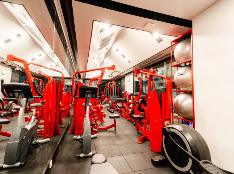 Red resistance machines line a modern gym with mirrored walls and a row of exercise balls on a red shelf.