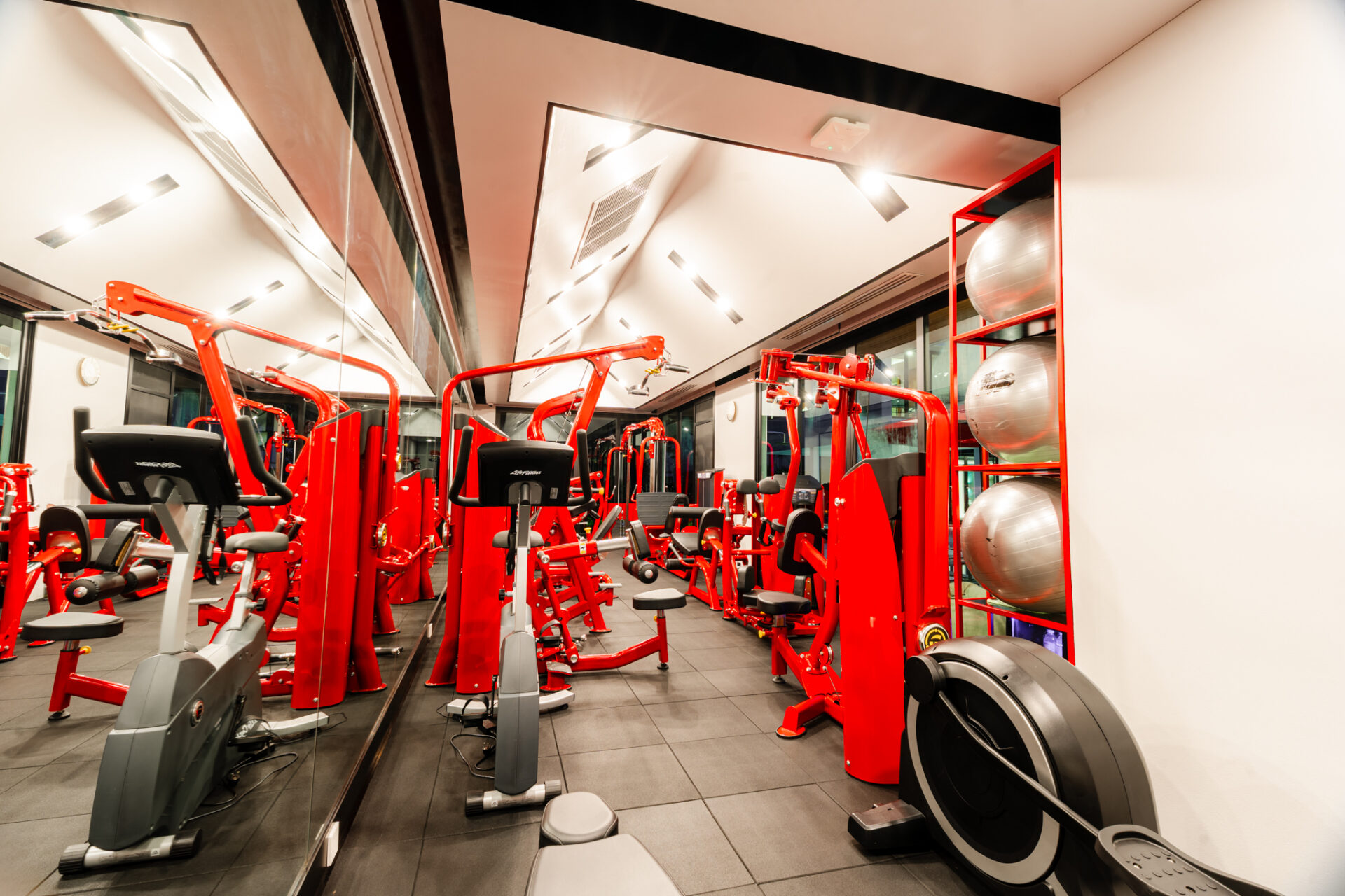 Red resistance machines line a modern gym with mirrored walls and a row of exercise balls on a red shelf.