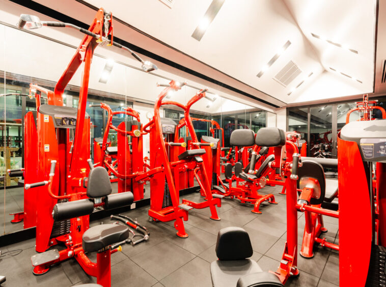 Red weight machines and benches in a bright gym with mirrored walls, reflecting the equipment.