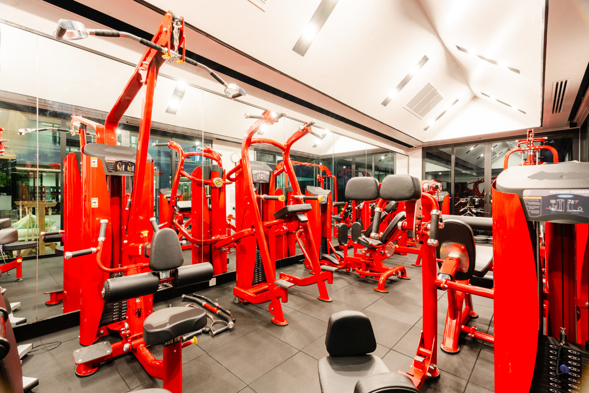 Red weight machines and benches in a bright gym with mirrored walls, reflecting the equipment.