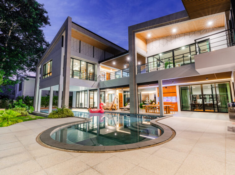 Modern two-story house with glass walls, outdoor lounge, and illuminated terrace around a curved swimming pool at dusk/night.