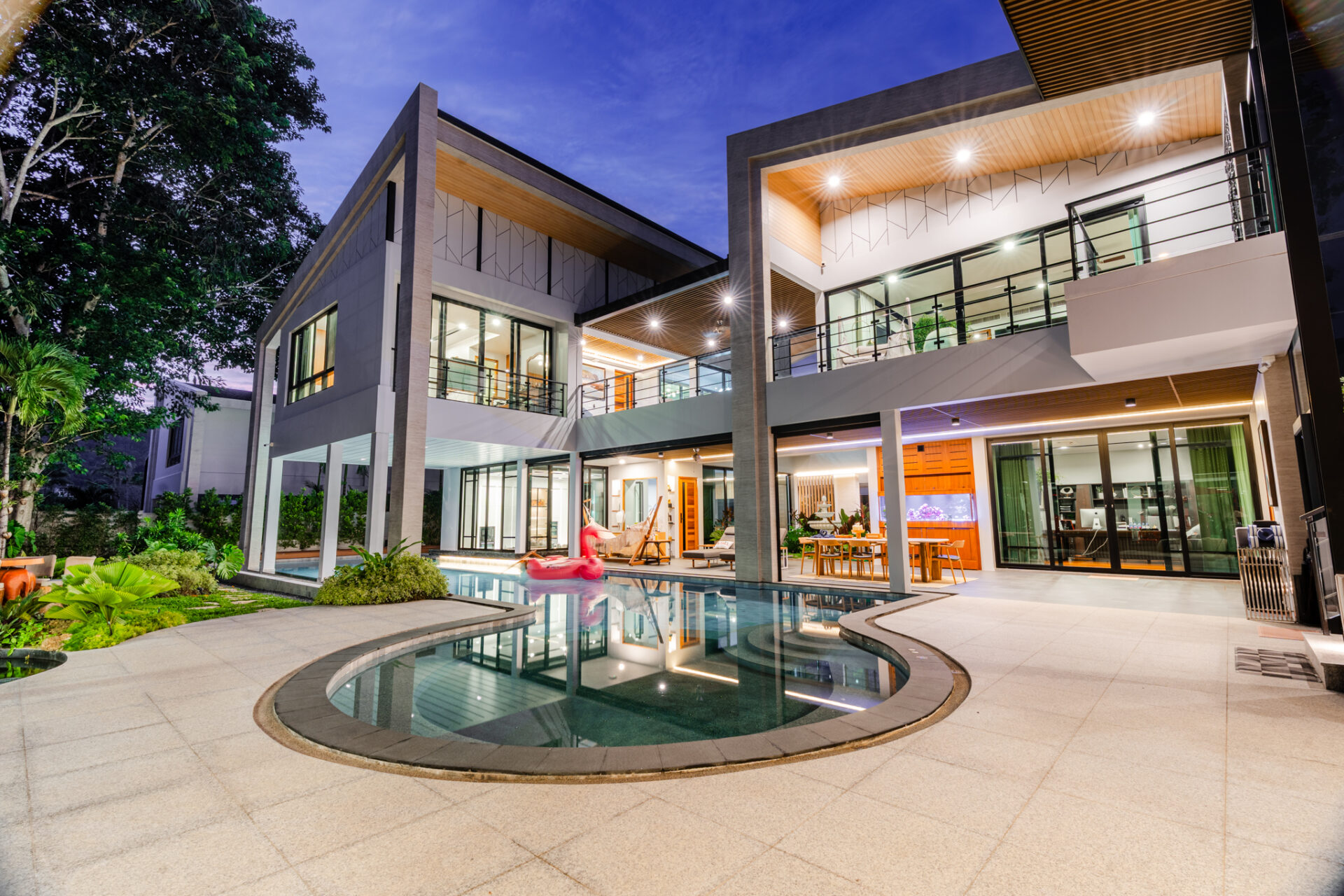 Modern two-story house with glass walls, outdoor lounge, and illuminated terrace around a curved swimming pool at dusk/night.