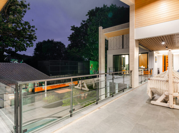Modern shaded balcony with glass railing, hammock, and outdoor seating overlooking a nighttime yard and trees.