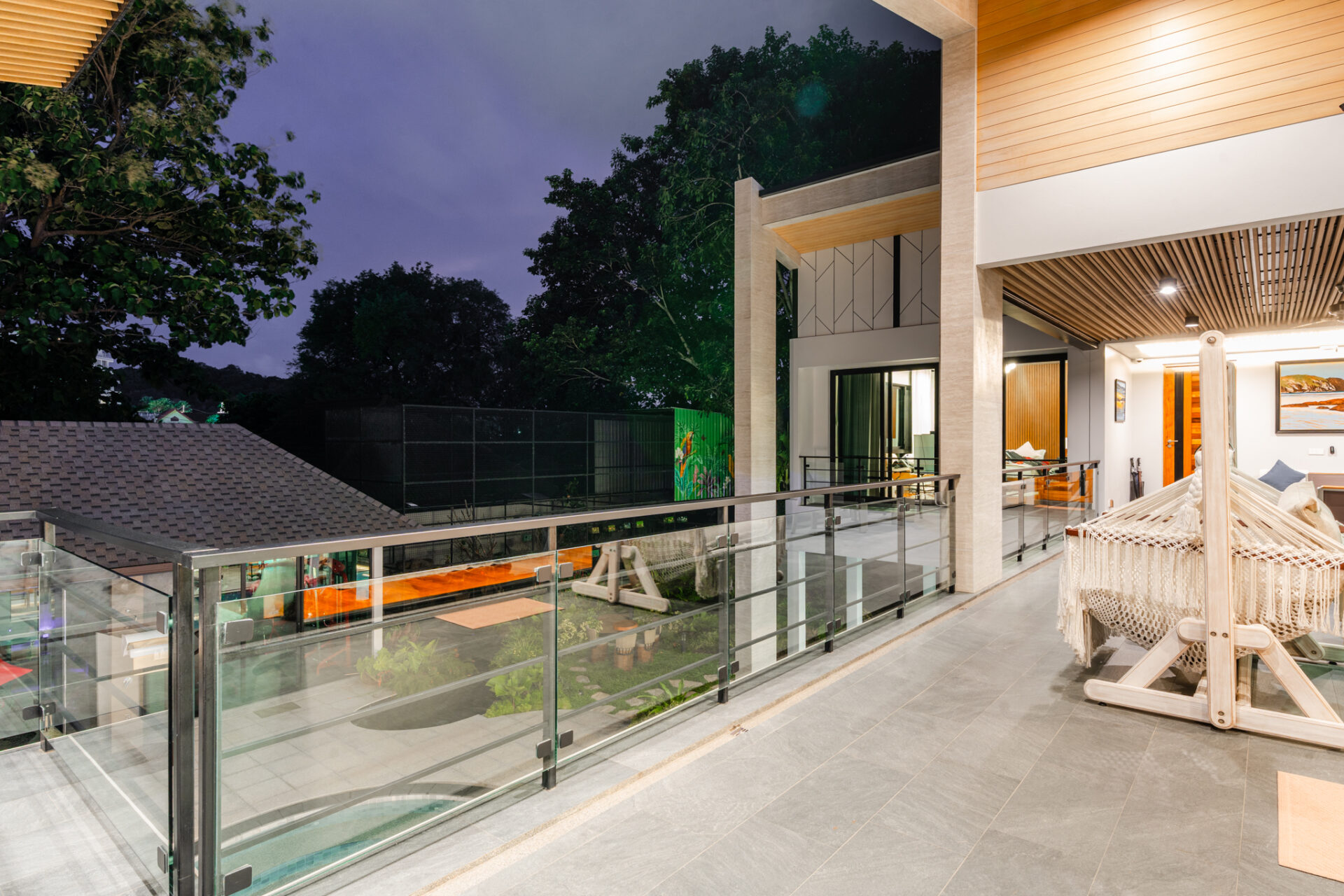 Modern shaded balcony with glass railing, hammock, and outdoor seating overlooking a nighttime yard and trees.