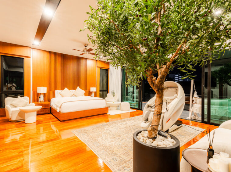 Luxurious hotel suite with a large indoor tree, warm wood paneling, and cream furnishings surrounding a plush bed and seating area at nightfall indoors.