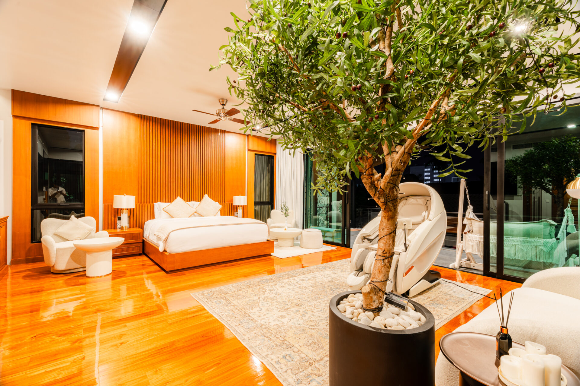 Luxurious hotel suite with a large indoor tree, warm wood paneling, and cream furnishings surrounding a plush bed and seating area at nightfall indoors.