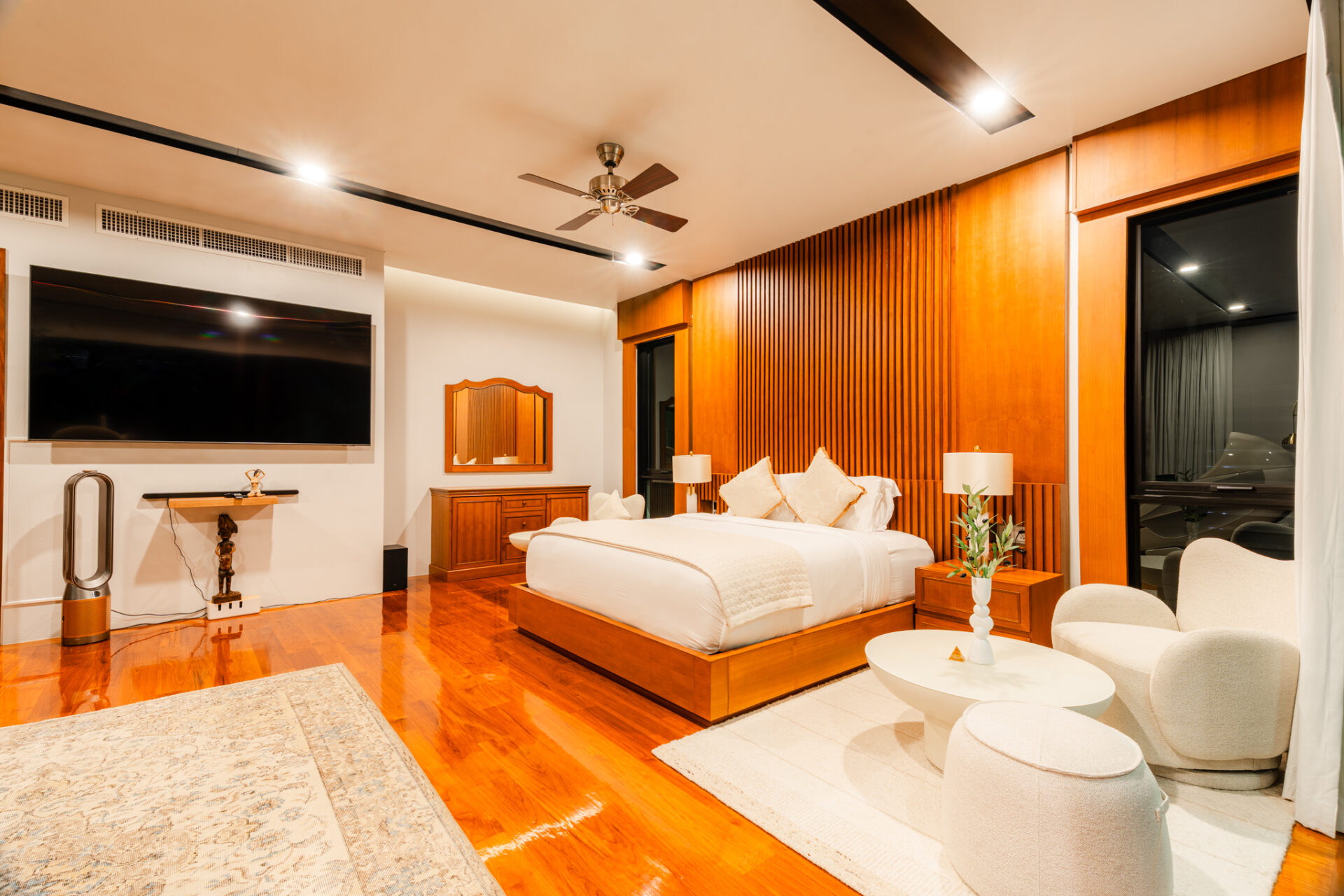 Luxurious hotel bedroom with a large bed, wood panel accents, and a wall-mounted flat-screen TV visible across the room.