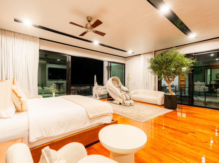 Luxurious hotel suite with white bedding, wooden floors, and a large potted tree by floor-to-ceiling windows at night