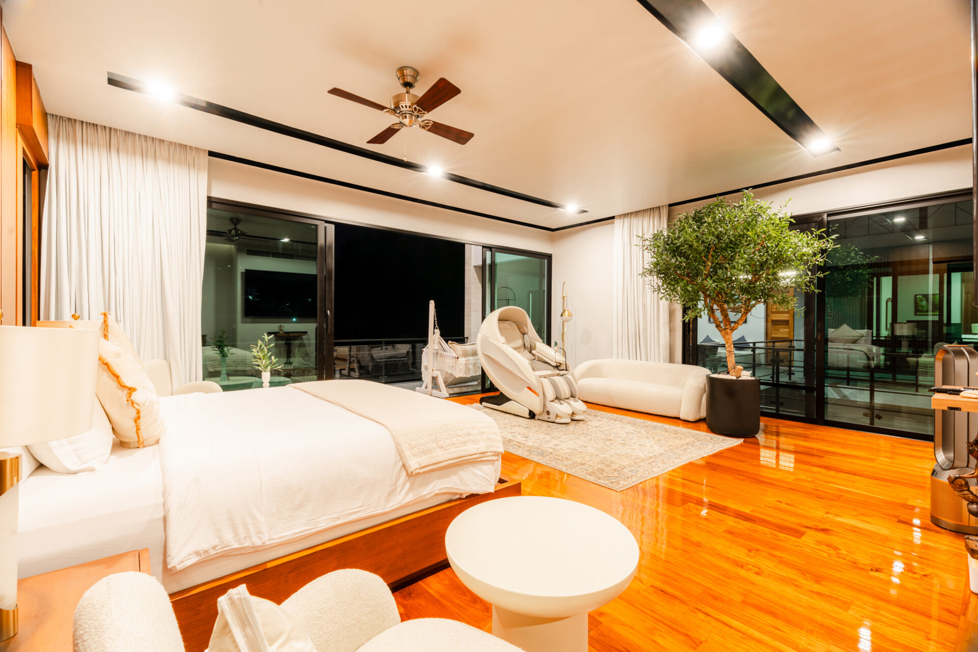 Luxurious hotel suite with white bedding, wooden floors, and a large potted tree by floor-to-ceiling windows at night