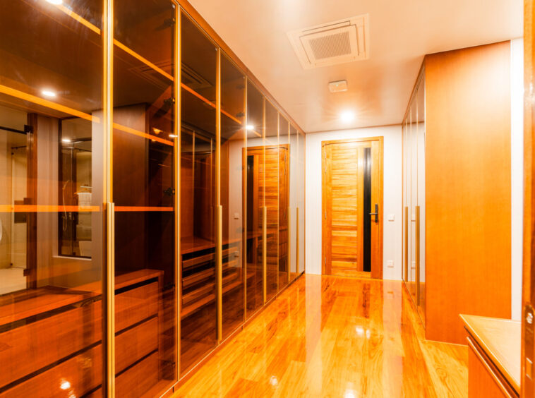 A modern walk-in closet with glass-front wardrobes along one side and wooden doors on the opposite wall, warm lighting.