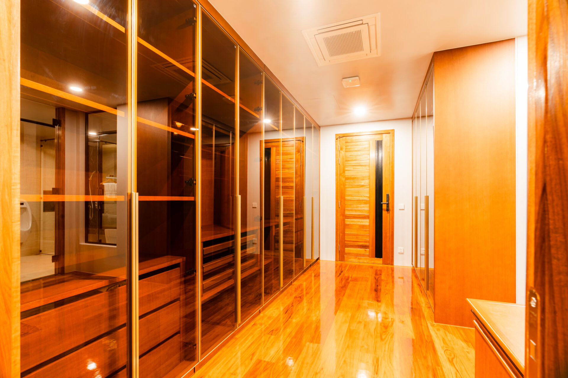 A modern walk-in closet with glass-front wardrobes along one side and wooden doors on the opposite wall, warm lighting.