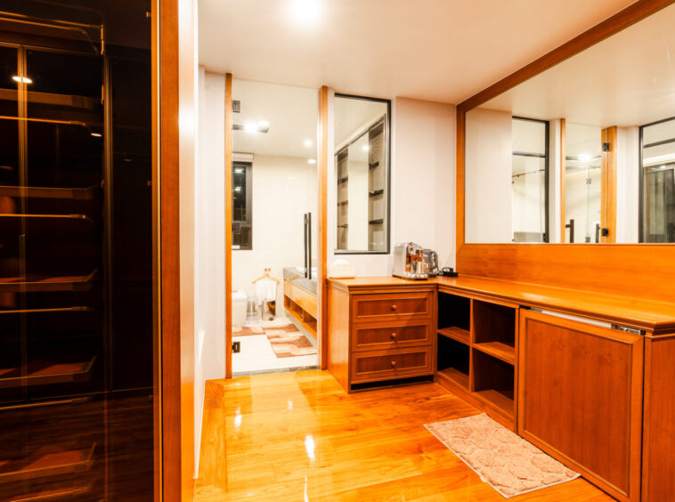 Wooden walk-in closet with large mirrors, drawers, and a coffee station; bathroom visible through a doorway.