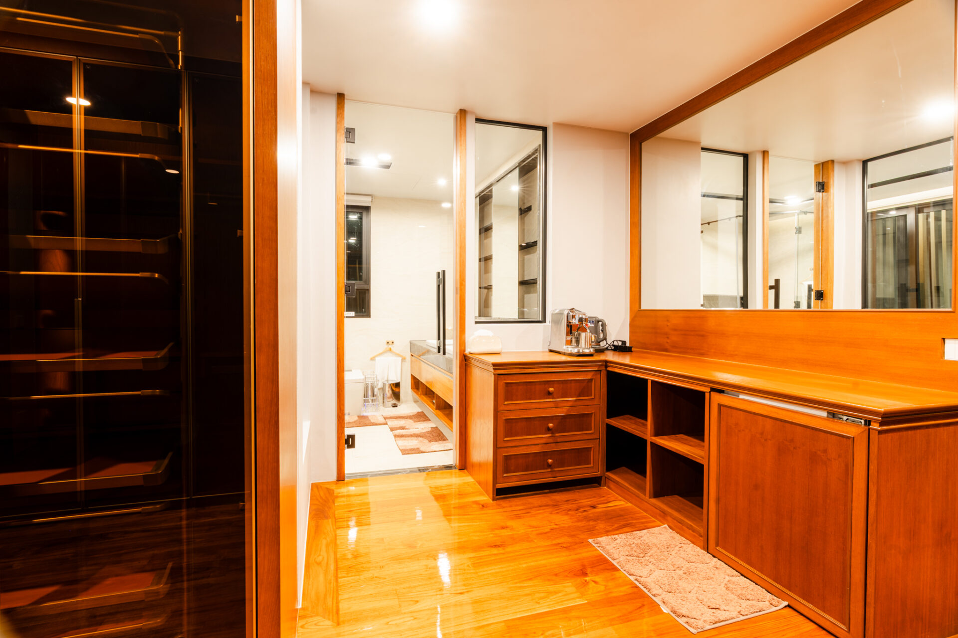 Wooden walk-in closet with large mirrors, drawers, and a coffee station; bathroom visible through a doorway.