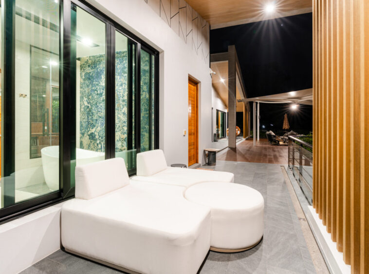 Night view of a modern outdoor patio with white cushioned seating, glass sliding doors, and a wooden slat wall under warm lighting on a balcony.