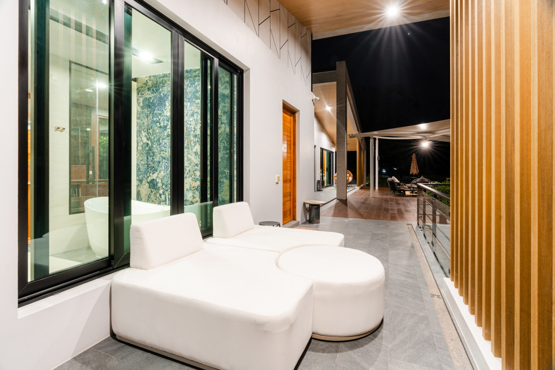 Night view of a modern outdoor patio with white cushioned seating, glass sliding doors, and a wooden slat wall under warm lighting on a balcony.