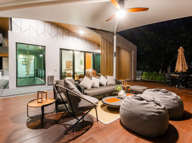 Nighttime outdoor lounge on a wooden deck with gray bean bags, cushioned seating, and wicker chairs beside glass doors to an illuminated interior.