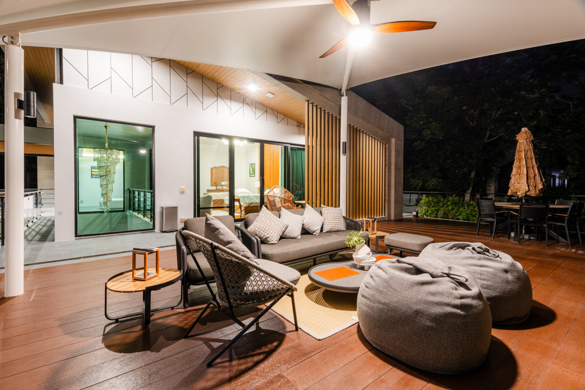 Nighttime outdoor lounge on a wooden deck with gray bean bags, cushioned seating, and wicker chairs beside glass doors to an illuminated interior.