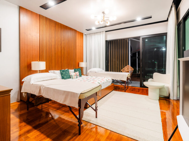 Hotel room with two beds, white bedding, teal pillows, and a wood-paneled wall; sliding glass doors open to a balcony outside.