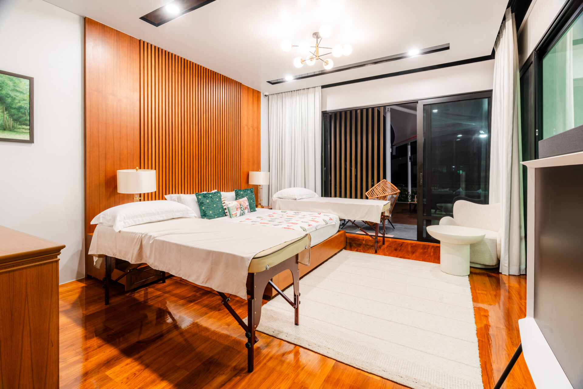 Hotel room with two beds, white bedding, teal pillows, and a wood-paneled wall; sliding glass doors open to a balcony outside.