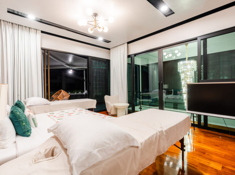 Bright hotel suite with multiple white beds, teal pillows, and a glossy wooden floor near large glass doors and a chandelier.