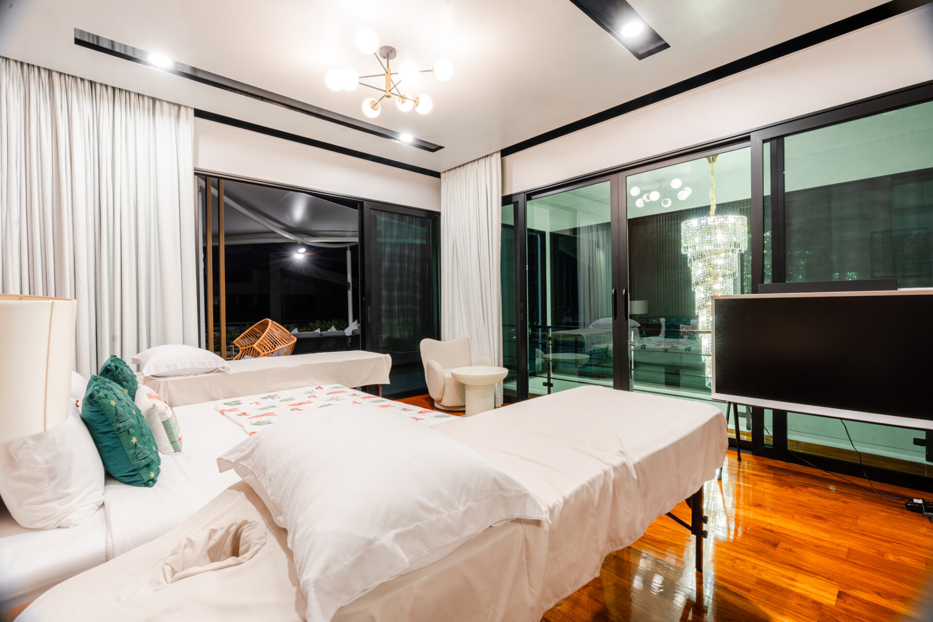 Bright hotel suite with multiple white beds, teal pillows, and a glossy wooden floor near large glass doors and a chandelier.