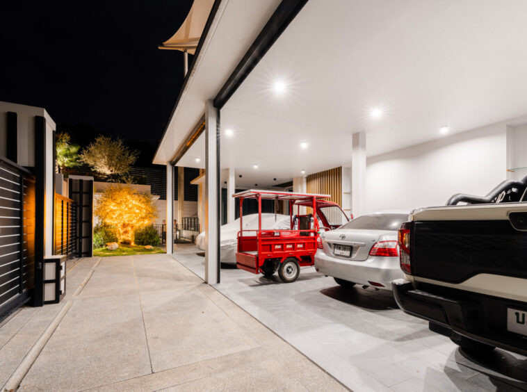 Nighttime view of a modern carport with bright ceiling lights, several parked cars, and a red utility cart in front of a white SUV/truck, exterior of a contemporary home