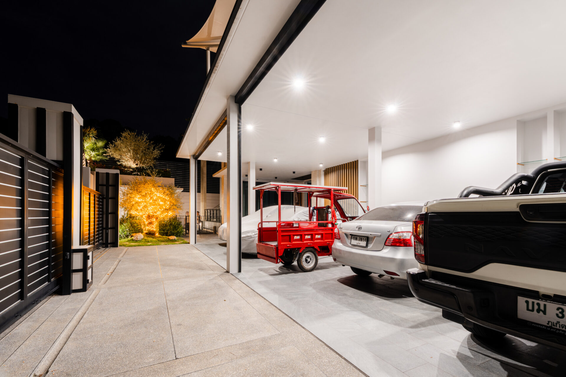 Nighttime view of a modern carport with bright ceiling lights, several parked cars, and a red utility cart in front of a white SUV/truck, exterior of a contemporary home
