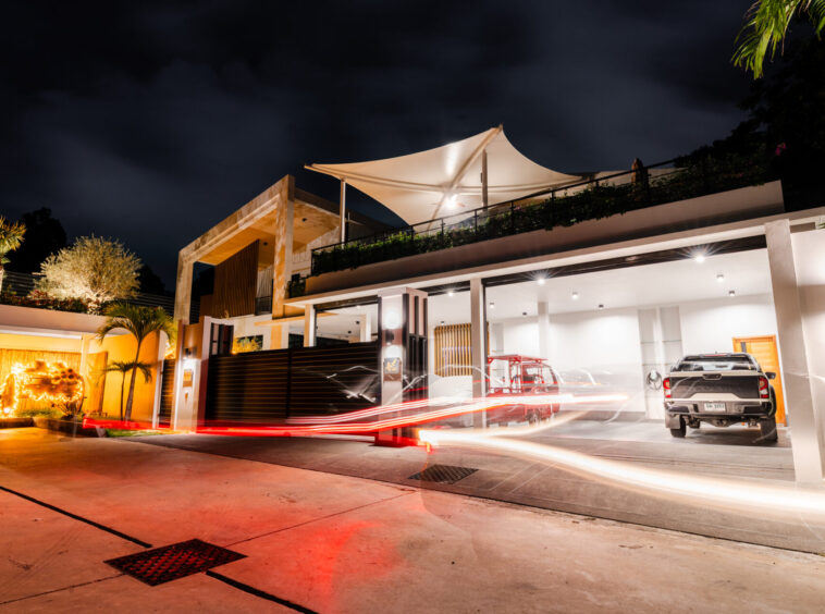 Nighttime exterior of a modern house with a white rooftop canopy, lit facade, and a parked SUV in the garage area.