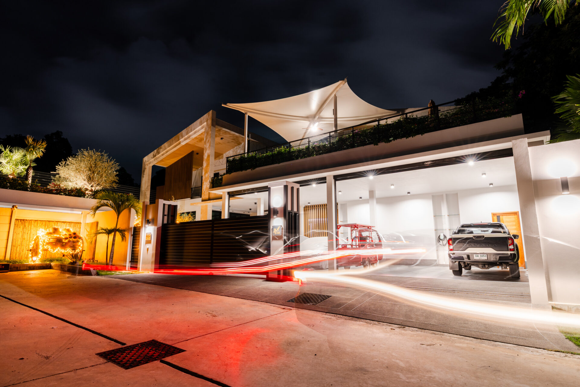 Nighttime exterior of a modern house with a white rooftop canopy, lit facade, and a parked SUV in the garage area.