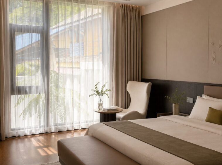 Bright hotel bedroom with large window and sheer curtains, a round side table, and a white armchair beside the bed.