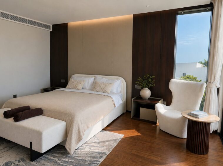A modern hotel-style bedroom with a large, beige bed, white pillows, and a cushioned bench at the foot; dark wood panels flank a beige headboard wall.