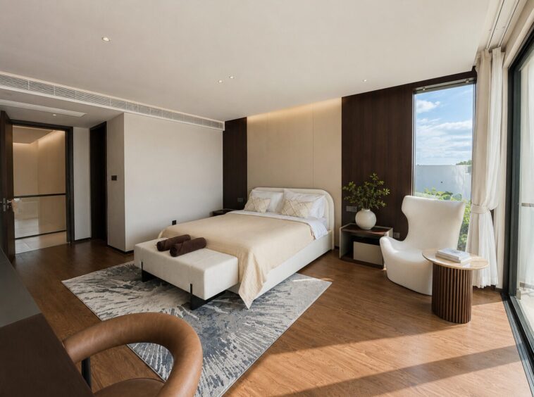 Modern hotel bedroom with a king bed, beige linens, bench at foot, and a wood-paneled wall beside a seating area by large windows.