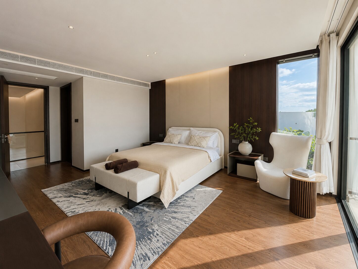 Modern hotel bedroom with a king bed, beige linens, bench at foot, and a wood-paneled wall beside a seating area by large windows.