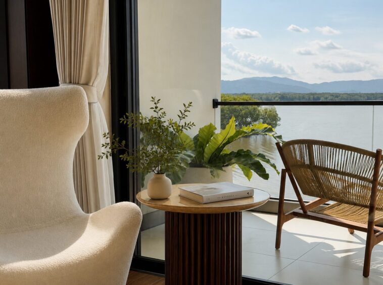 Cream upholstery chair and a woven wicker chair beside a round wooden table with a vase of greenery and books, near a glass balcony door with a lake view beyond.