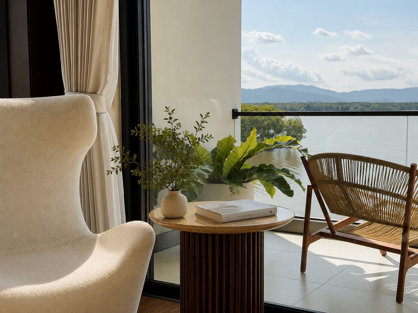 Cream upholstery chair and a woven wicker chair beside a round wooden table with a vase of greenery and books, near a glass balcony door with a lake view beyond.