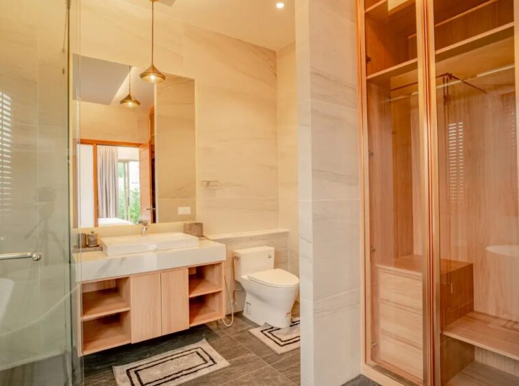 Modern bathroom with a glass shower on the left, a light wood vanity, and a toilet with a wooden sauna cabinet on the right.