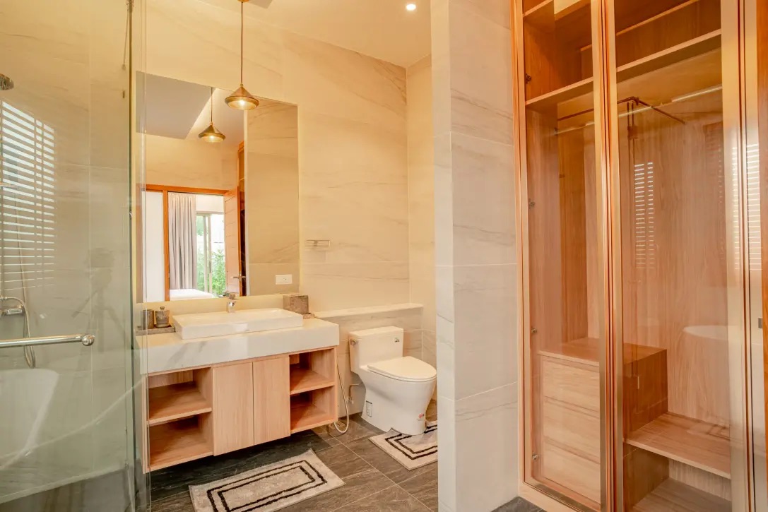Modern bathroom with a glass shower on the left, a light wood vanity, and a toilet with a wooden sauna cabinet on the right.