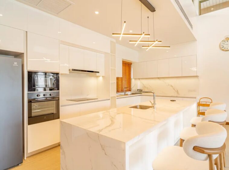 Bright white modern kitchen with marble island and countertops, stainless steel appliances, and pendant lighting; white bar stools line the island edge as a breakfast seating area.
