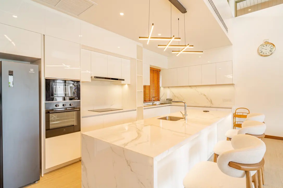 Bright white modern kitchen with marble island and countertops, stainless steel appliances, and pendant lighting; white bar stools line the island edge as a breakfast seating area.