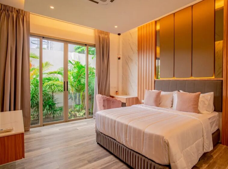 Modern hotel room with a king bed, cream bedding and pink pillows, wooden panels, marble accent wall, and sliding glass doors to a green garden outside.