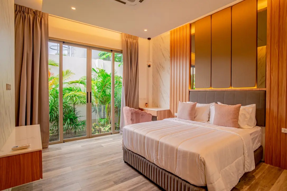 Modern hotel room with a king bed, cream bedding and pink pillows, wooden panels, marble accent wall, and sliding glass doors to a green garden outside.