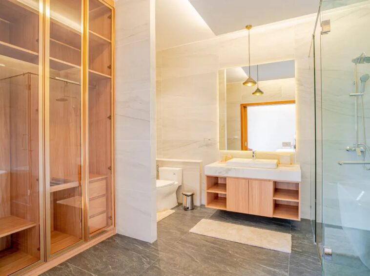 Modern bathroom with glass-walled shower, wooden vanity, and large mirror reflecting a doorway; neutral tones and marble accents present.