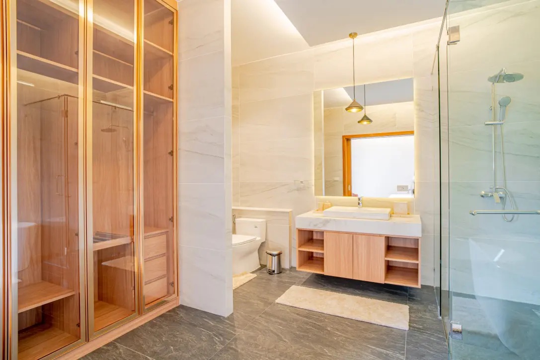 Modern bathroom with glass-walled shower, wooden vanity, and large mirror reflecting a doorway; neutral tones and marble accents present.