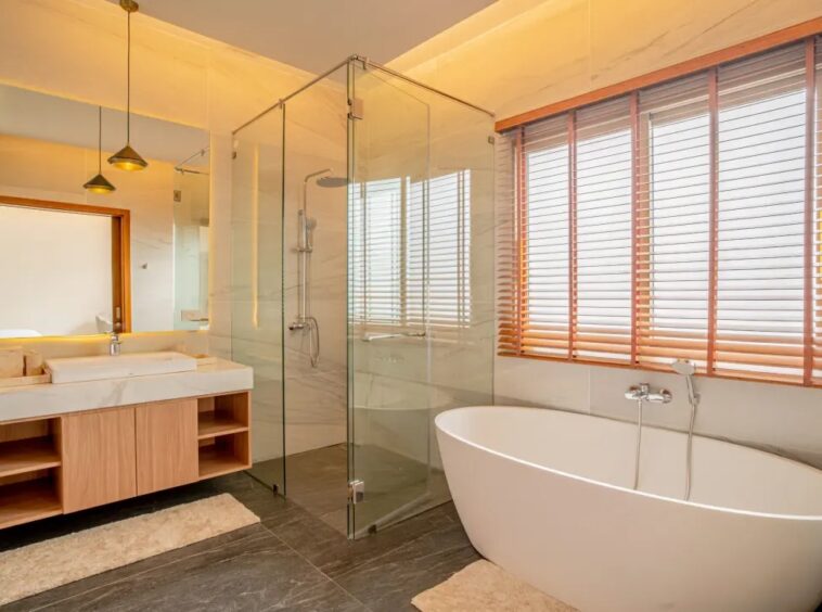 Modern bathroom with a glass-enclosed shower, freestanding oval tub by a large window with wood blinds, and a wooden vanity under warm lighting.