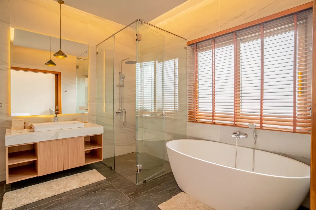 Modern bathroom with a glass-enclosed shower, freestanding oval tub by a large window with wood blinds, and a wooden vanity under warm lighting.