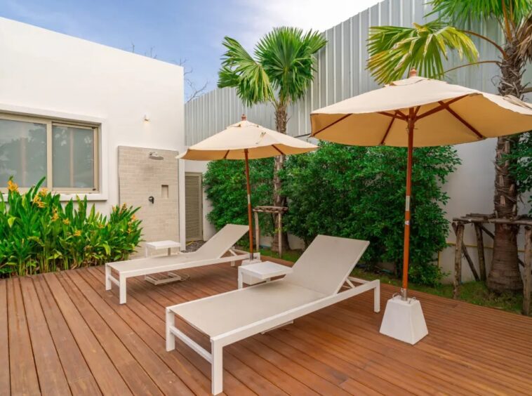 Outdoor wooden deck with two beige lounge chairs under large beige patio umbrellas, bordered by tropical plants and a white building wall.