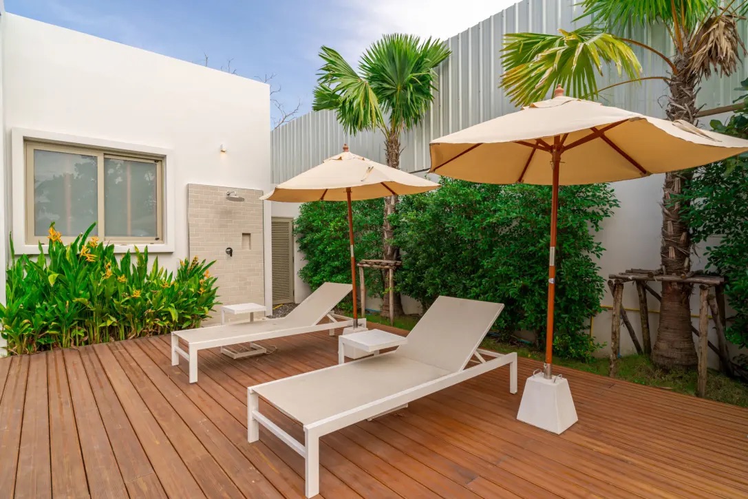 Outdoor wooden deck with two beige lounge chairs under large beige patio umbrellas, bordered by tropical plants and a white building wall.