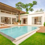 Modern white villa with a rectangular pool, wooden deck, and open living spaces by glass doors right outside the home.