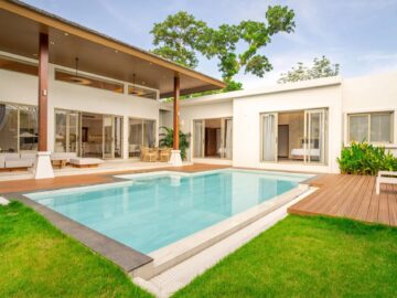 Modern white villa with a rectangular pool, wooden deck, and open living spaces by glass doors right outside the home.
