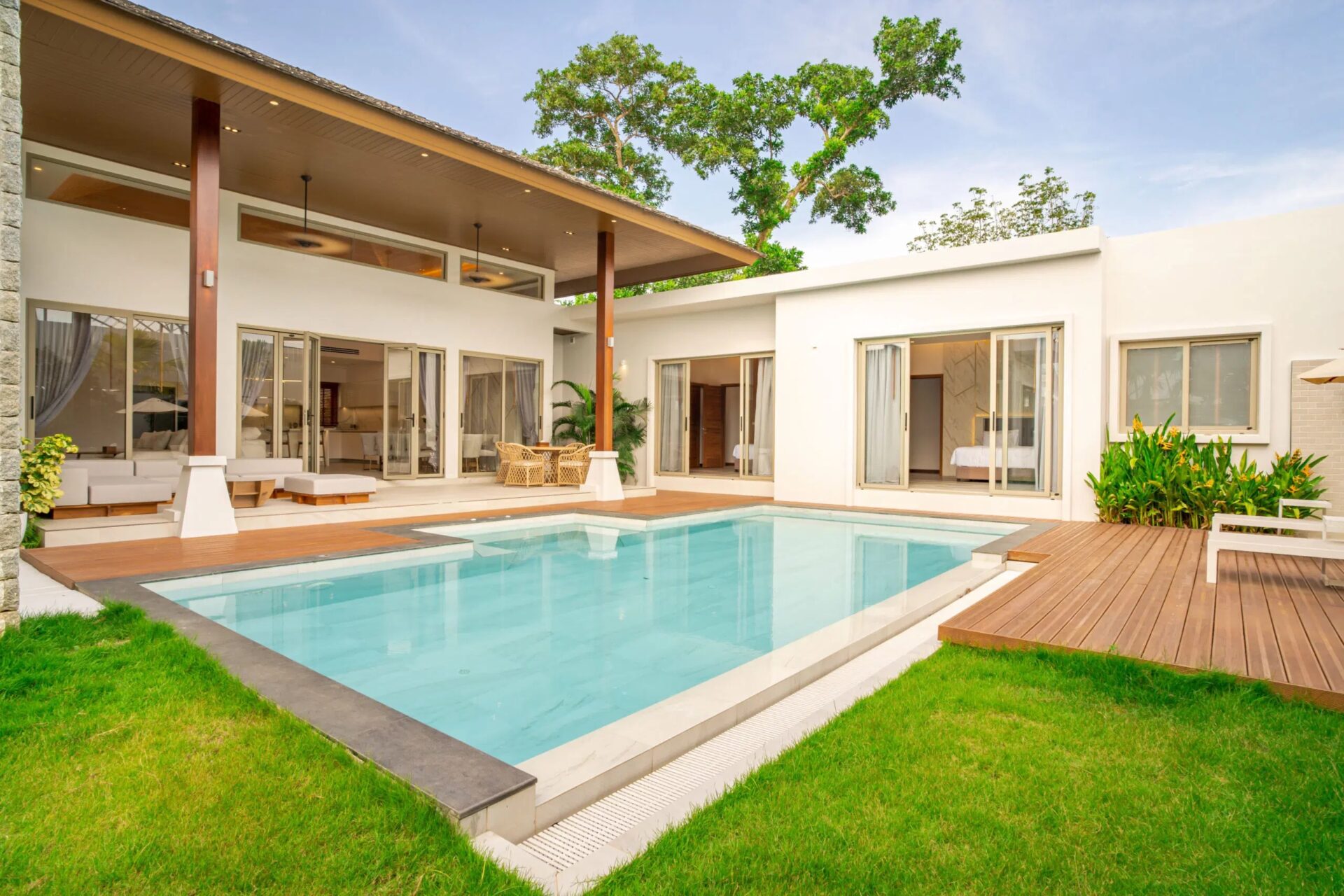 Modern white villa with a rectangular pool, wooden deck, and open living spaces by glass doors right outside the home.